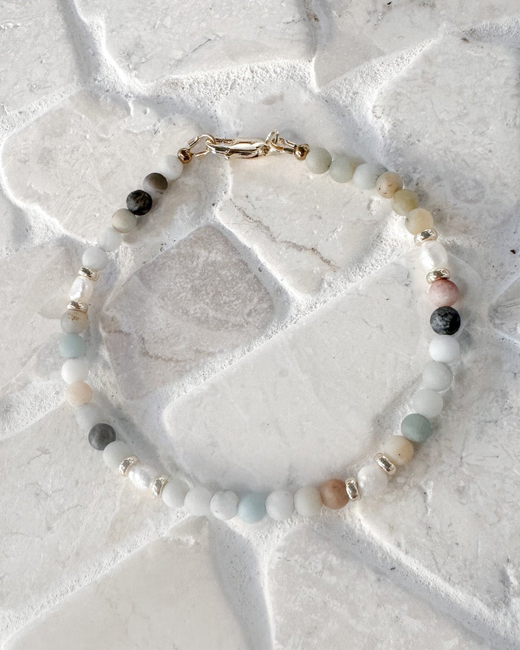 A delicate beaded bracelet with a gold clasp, featuring a mix of light grey, white, pale blue, and dark grey amazonite beads, laid flat on a textured white stone surface.