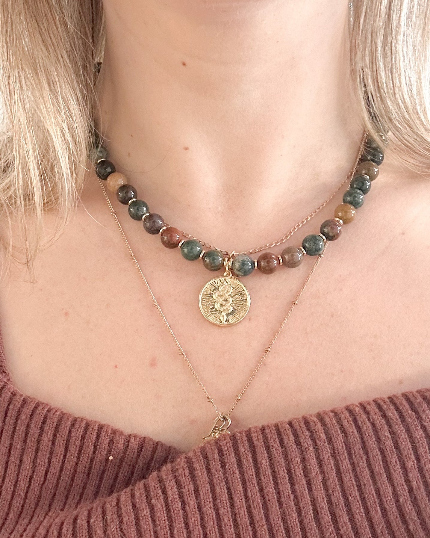 A close-up of a model wearing a layered necklace set. The top layer is a bohemian-style beaded necklace with green, brown, and grey indian agate gemstones, featuring a gold-toned sunburst snake medallion.
