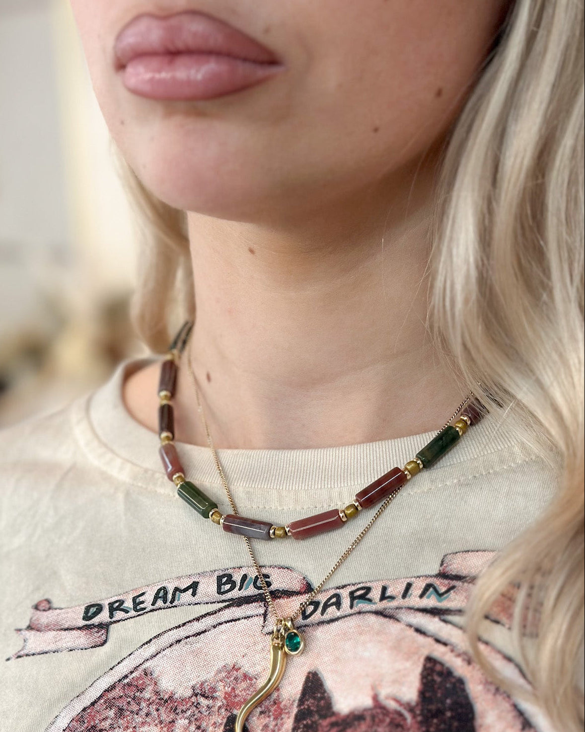 A close-up lifestyle shot of a woman wearing an Indian agate beaded necklace with gold-toned chains and a small green-and-gold charm, paired with a graphic t-shirt.