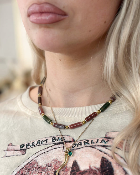 A close-up lifestyle shot of a woman wearing an Indian agate beaded necklace with gold-toned chains and a small green-and-gold charm, paired with a graphic t-shirt.