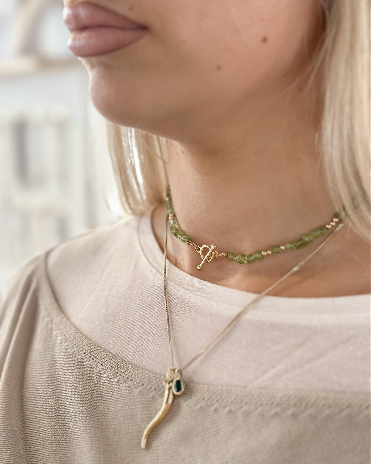 Close-up of a woman wearing layered necklaces, including a green beaded choker with a gold toggle clasp and a gold cornicello chilli pendant necklace with a small green stone.