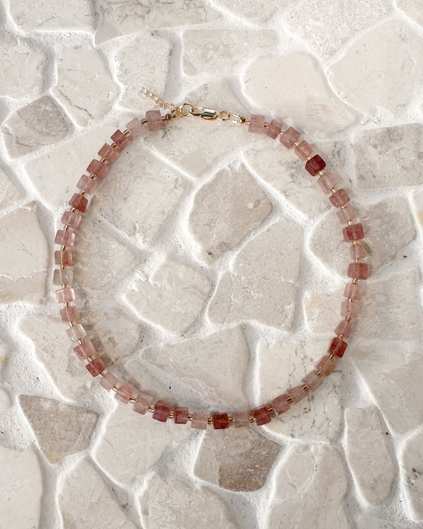 A beaded necklace made of small faceted cube strawberry quartz gemstones with a gold clasp, displayed on a light beige stone-tile surface.