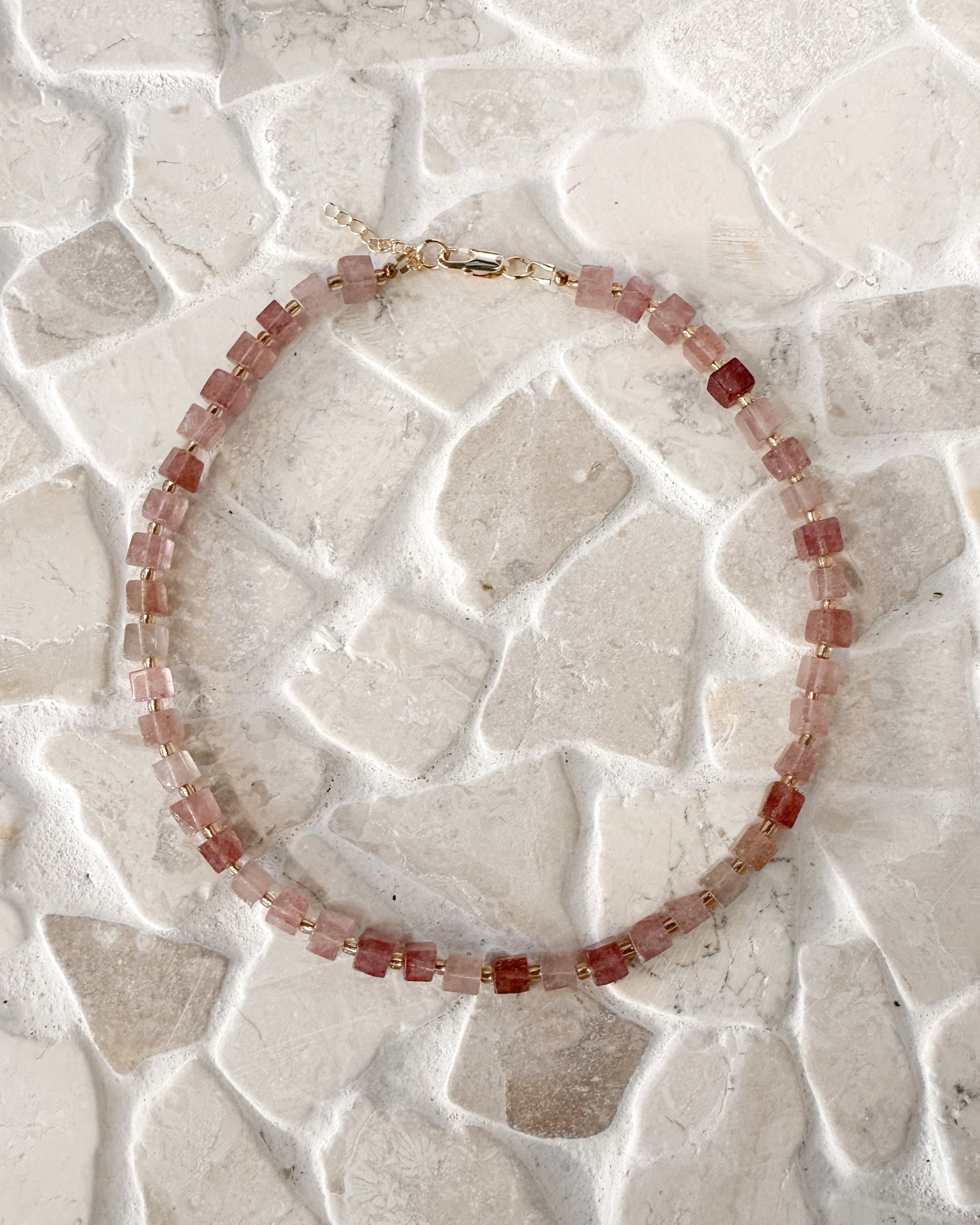 A beaded necklace made of small faceted cube strawberry quartz gemstones with a gold clasp, displayed on a light beige stone-tile surface.
