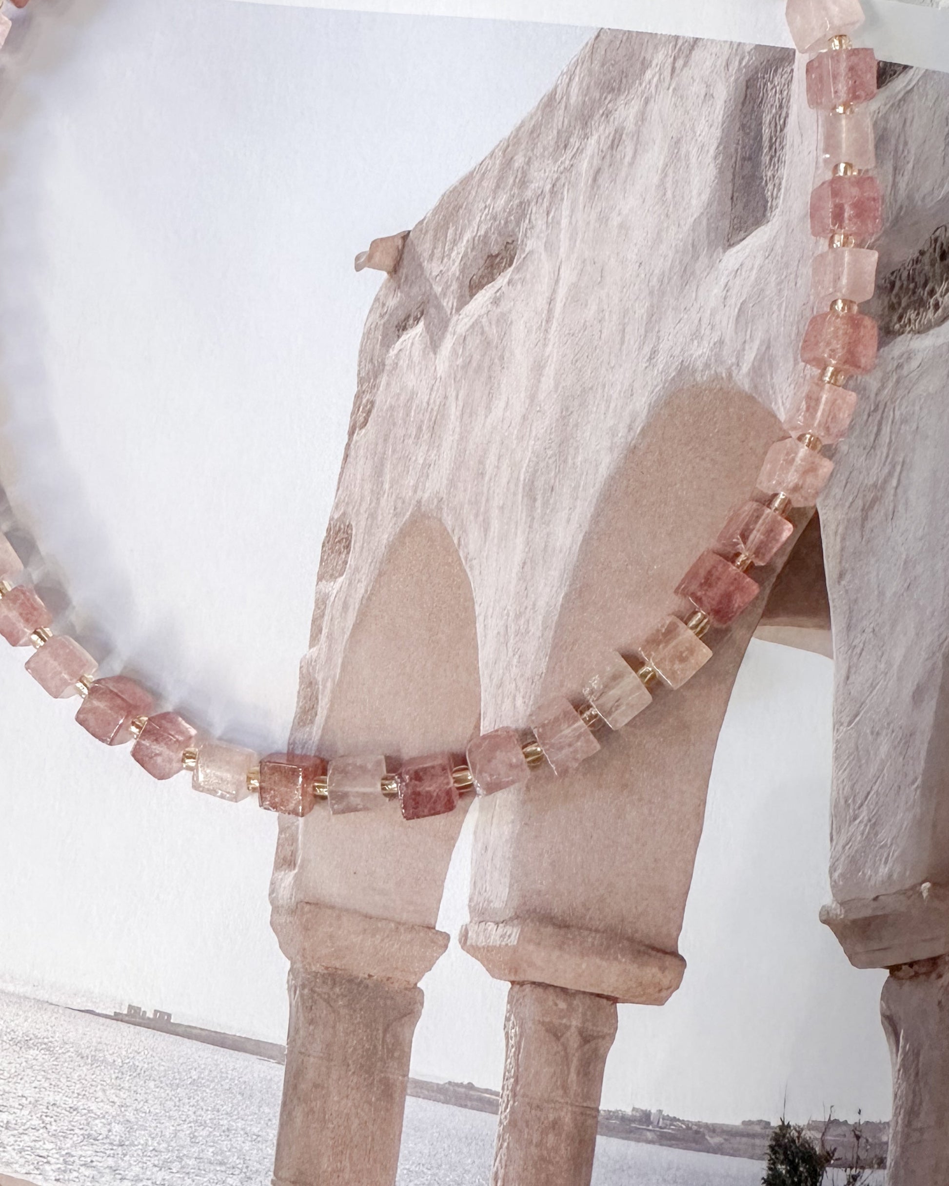 A beaded necklace made of strawberry quartz faceted square beads draped over an open book showing a coastal architectural scene.
