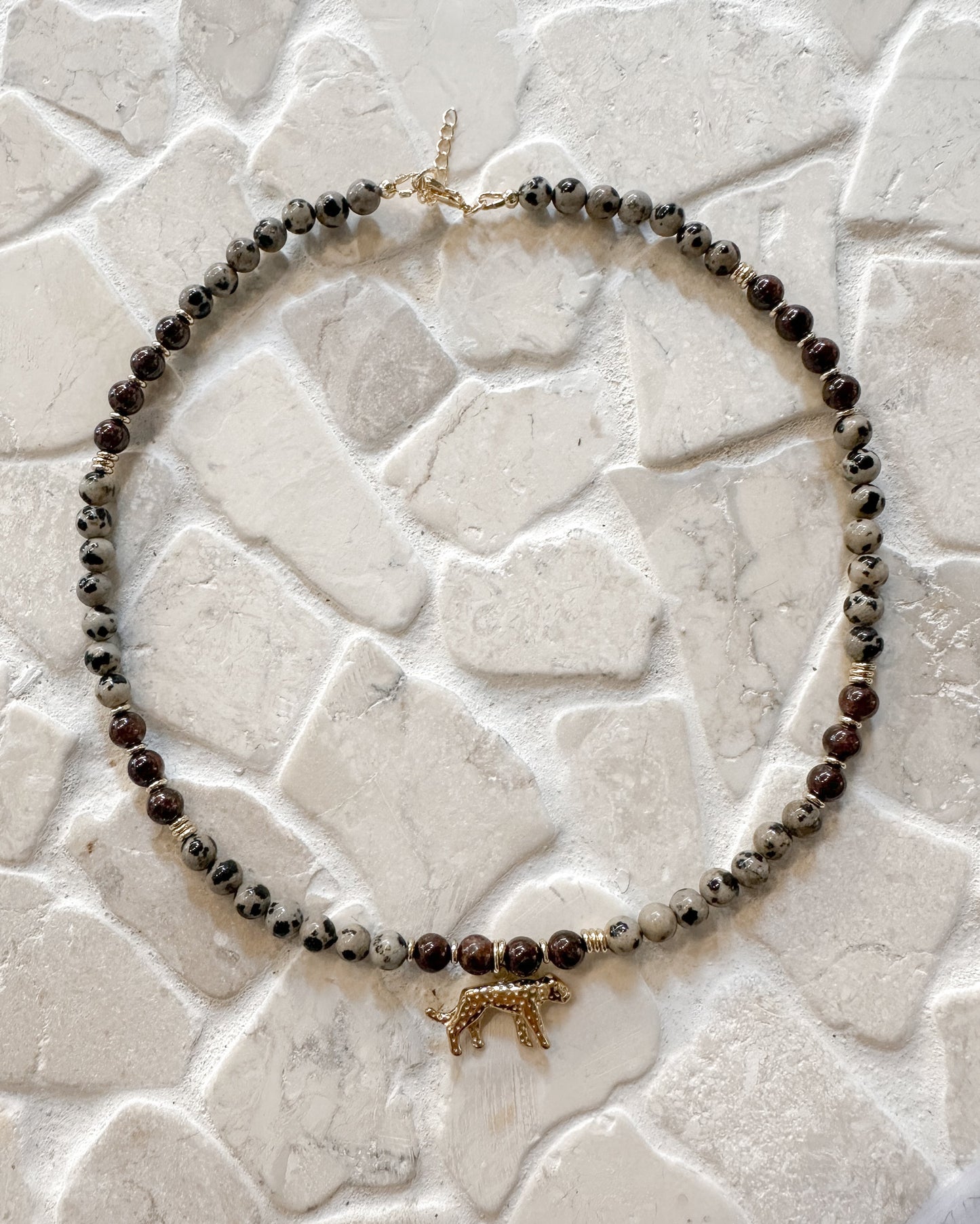 A beaded necklace with dark red garnet and Dalmatian jasper marbled beads, featuring a small gold leopard charm in the center, displayed on a textured white stone surface.