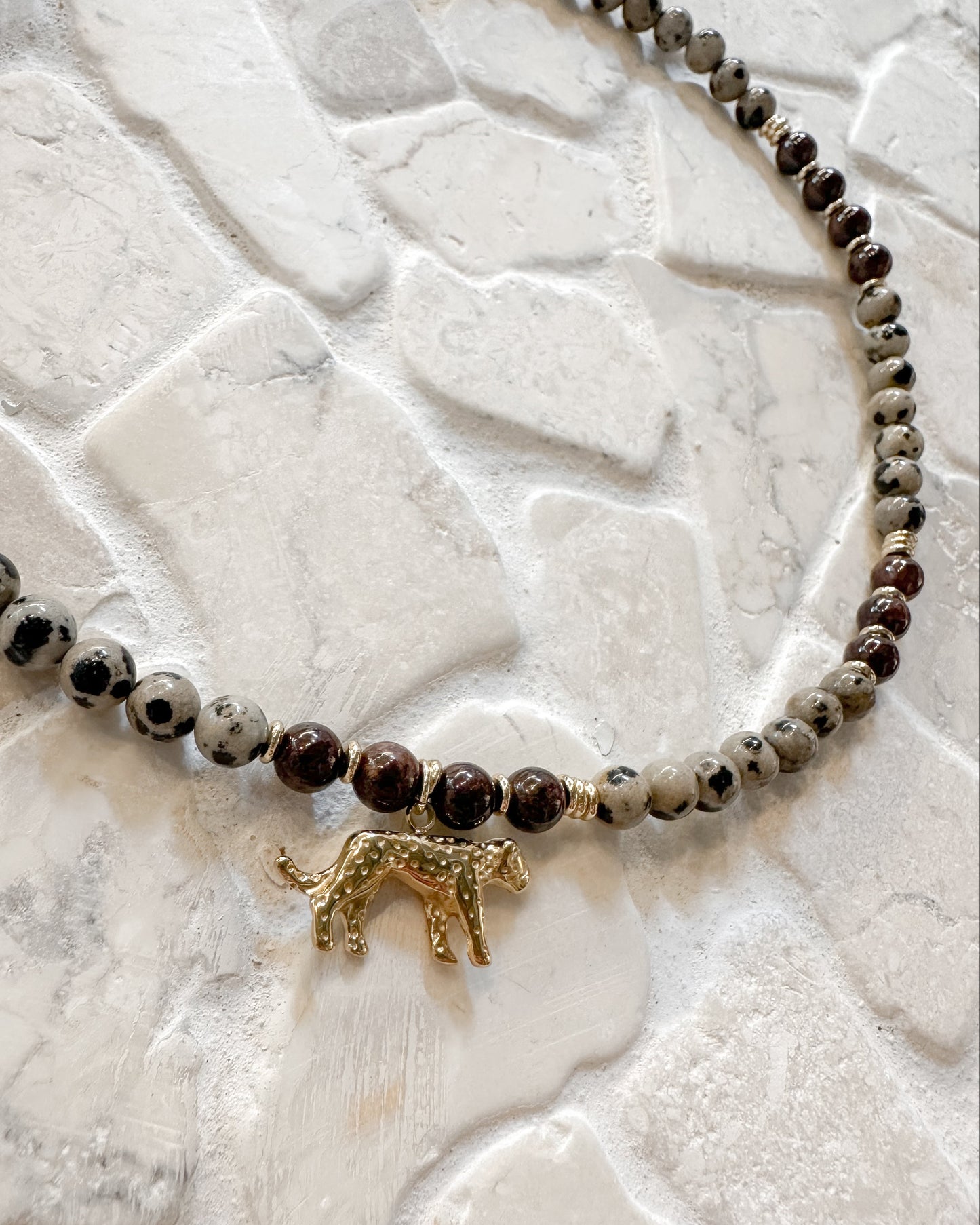 A beaded necklace featuring dark brown and black spotted beads, light gray and white marbled beads, and a small gold leopard charm, displayed on a textured white stone surface.