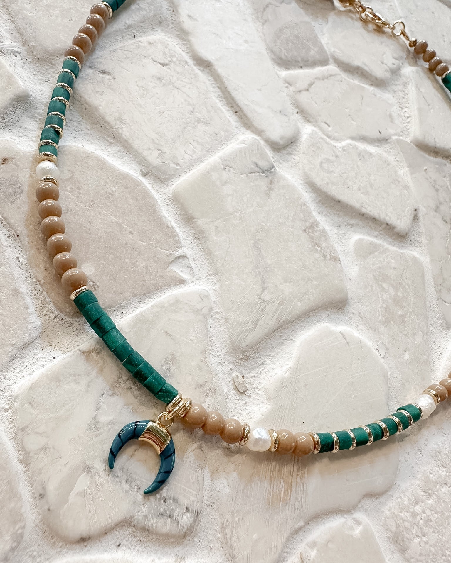 A bohemian-style beaded necklace with turquoise green and beige beads, white pearl accents, and a crescent moon pendant, displayed on a textured white stone surface.