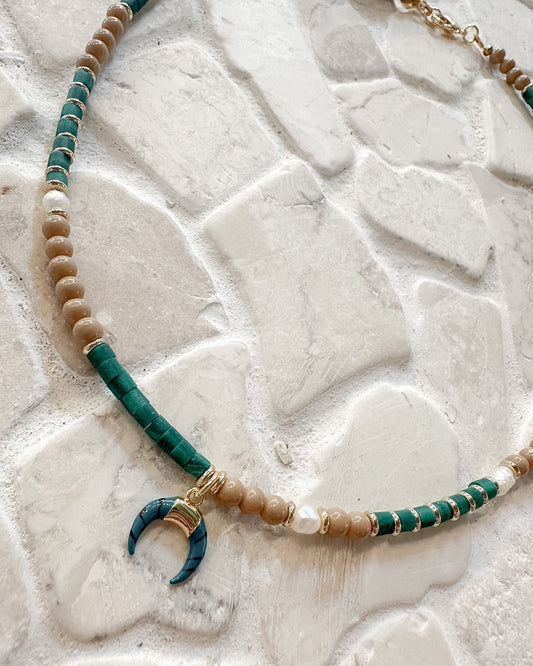 A bohemian-style beaded necklace with turquoise green and beige beads, white pearl accents, and a crescent moon pendant, displayed on a textured white stone surface.