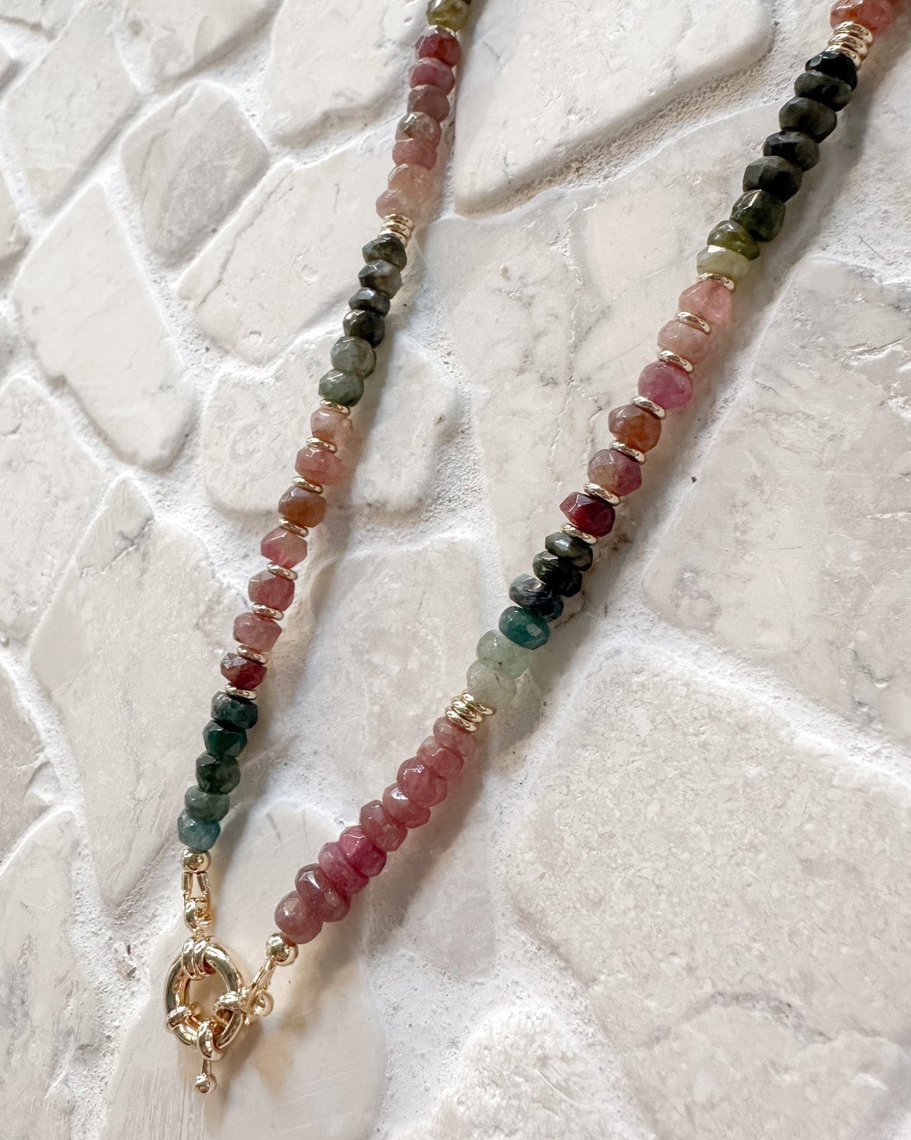 A beaded tourmaline necklace with faceted gemstone beads in pink, green, red, and black, accented with small gold spacer rings and a gold lobster clasp, displayed on a textured white surface.
