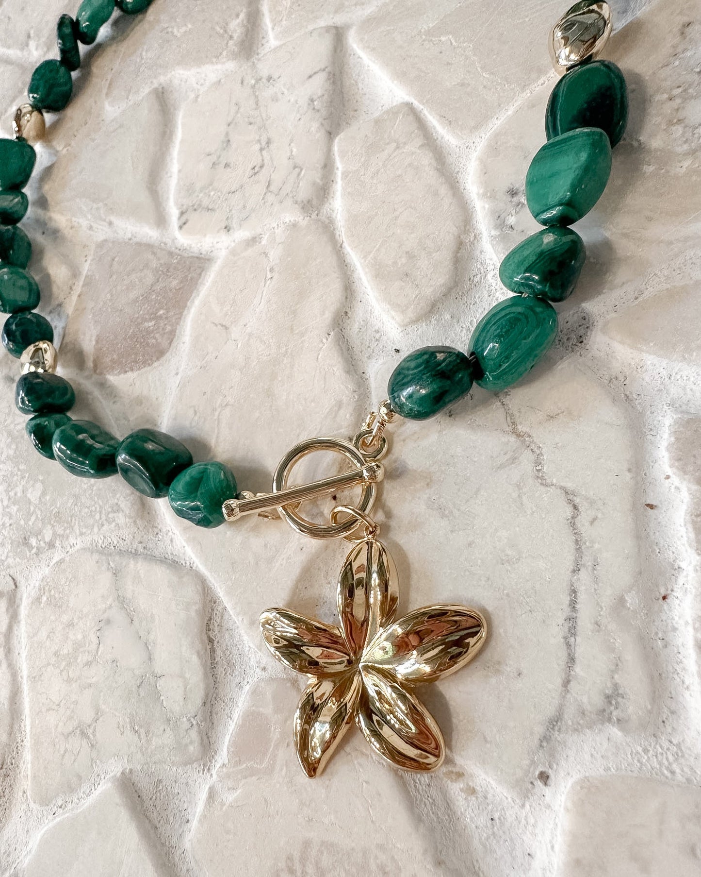 A beaded necklace featuring polished green malachite beads and a shiny gold flower-shaped pendant, displayed on a textured light beige stone surface.