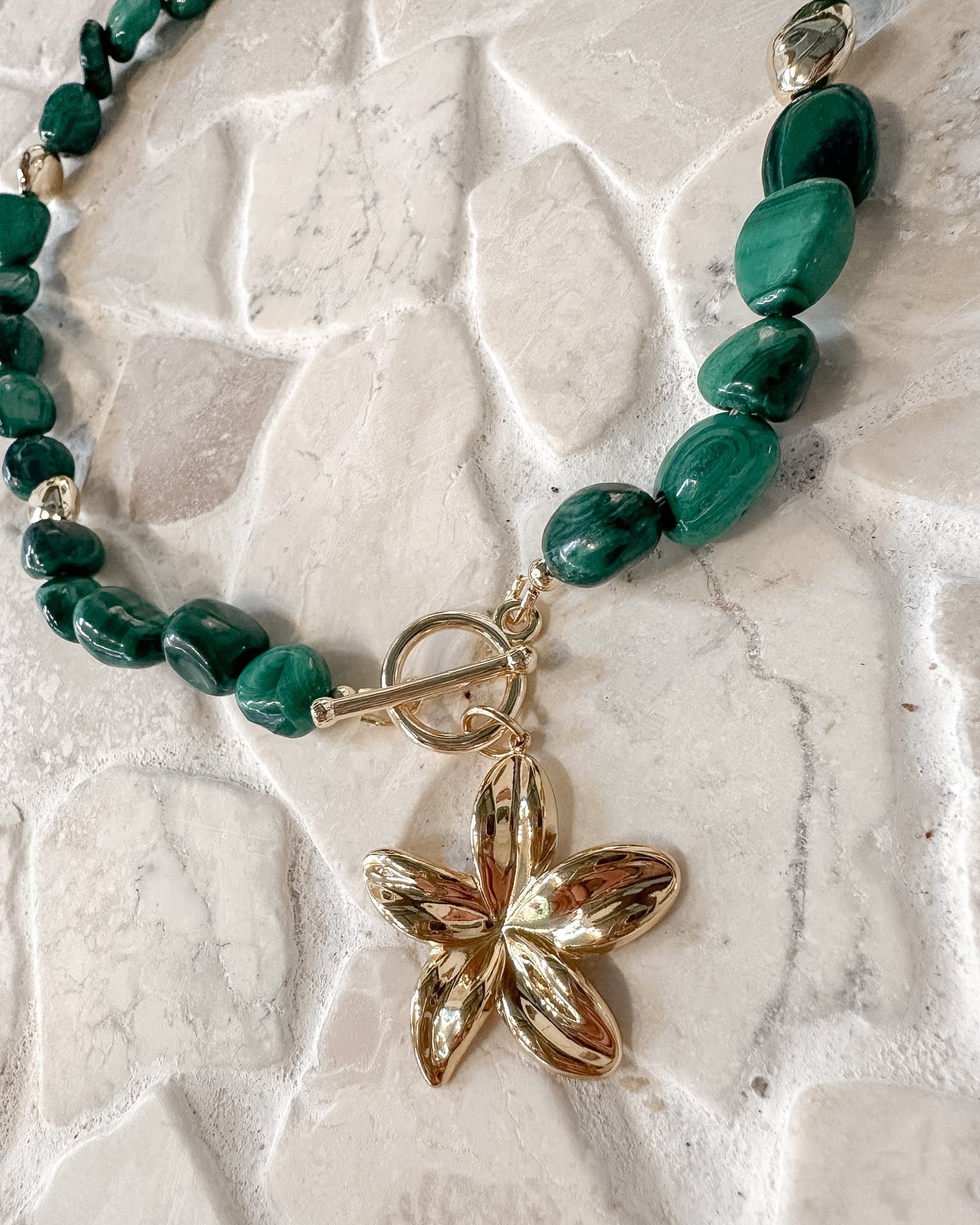 A beaded necklace featuring polished green malachite beads and a shiny gold flower-shaped pendant, displayed on a textured light beige stone surface.